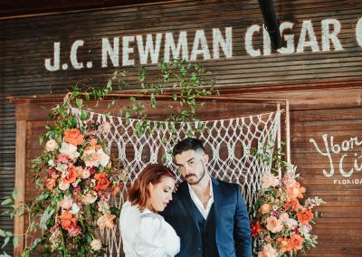 bride and groom holding cigars in front of beadboard and flowers in cigar factory