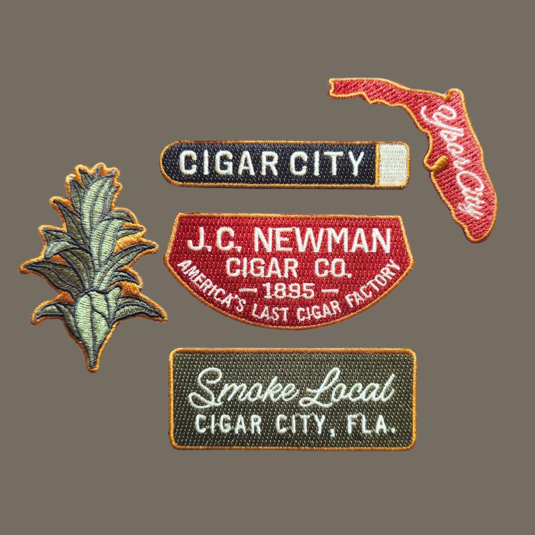 a variety of embroidered patches: a tobacco leaf, cigar city sign, state of florida, J.C. Newman Cigar co. and smoke local sign