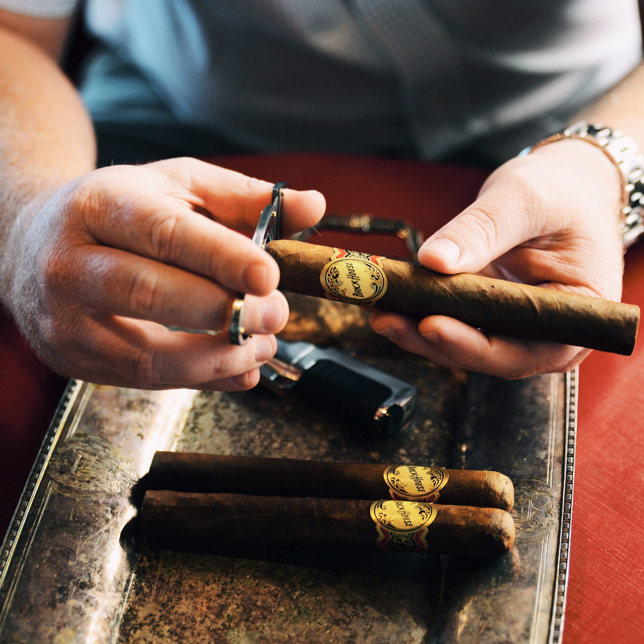hands cutting a brick house cigar over an ashtray with two other uncut brick house cigars