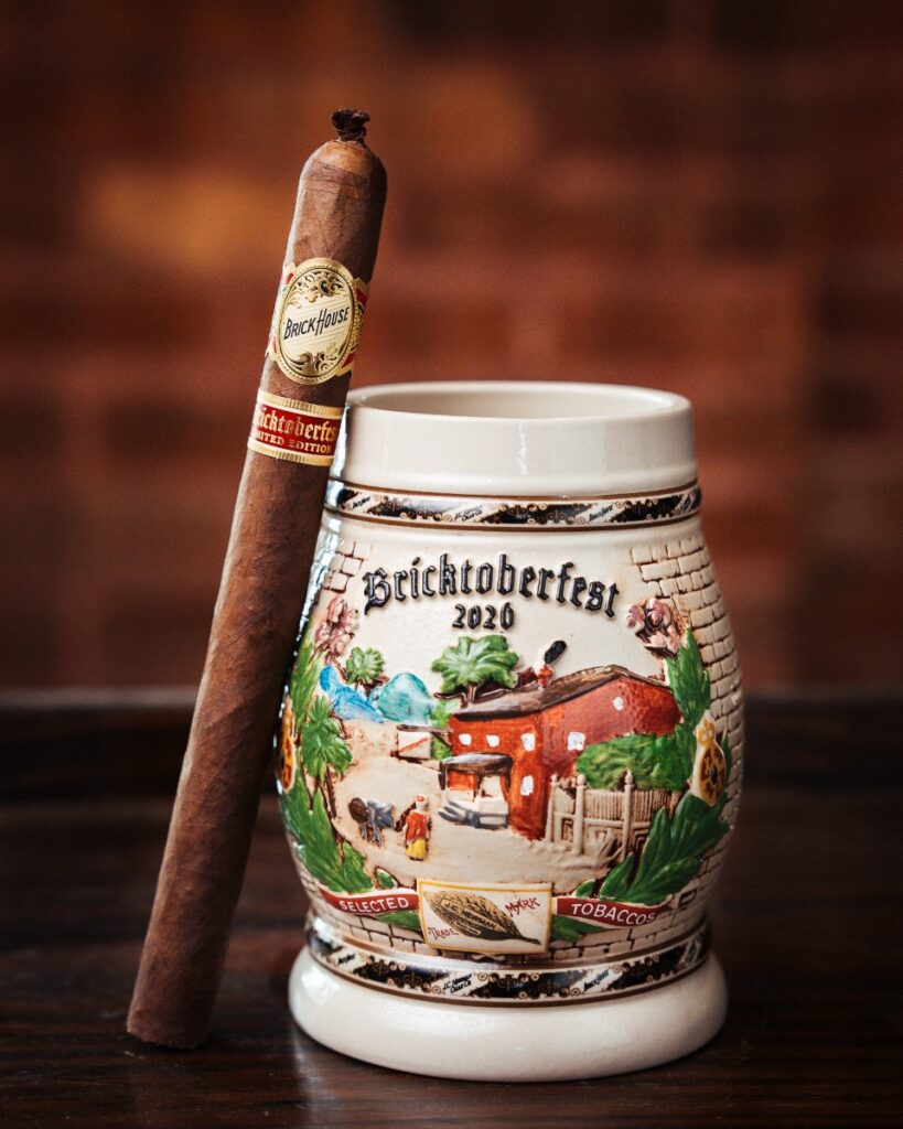 Single Brick House Bricktoberfest Cigar with pigtail leaned against the 2026 Brick House Bricktoberfest Beer Stein that features a vista of the Brick House Cigar box interior
